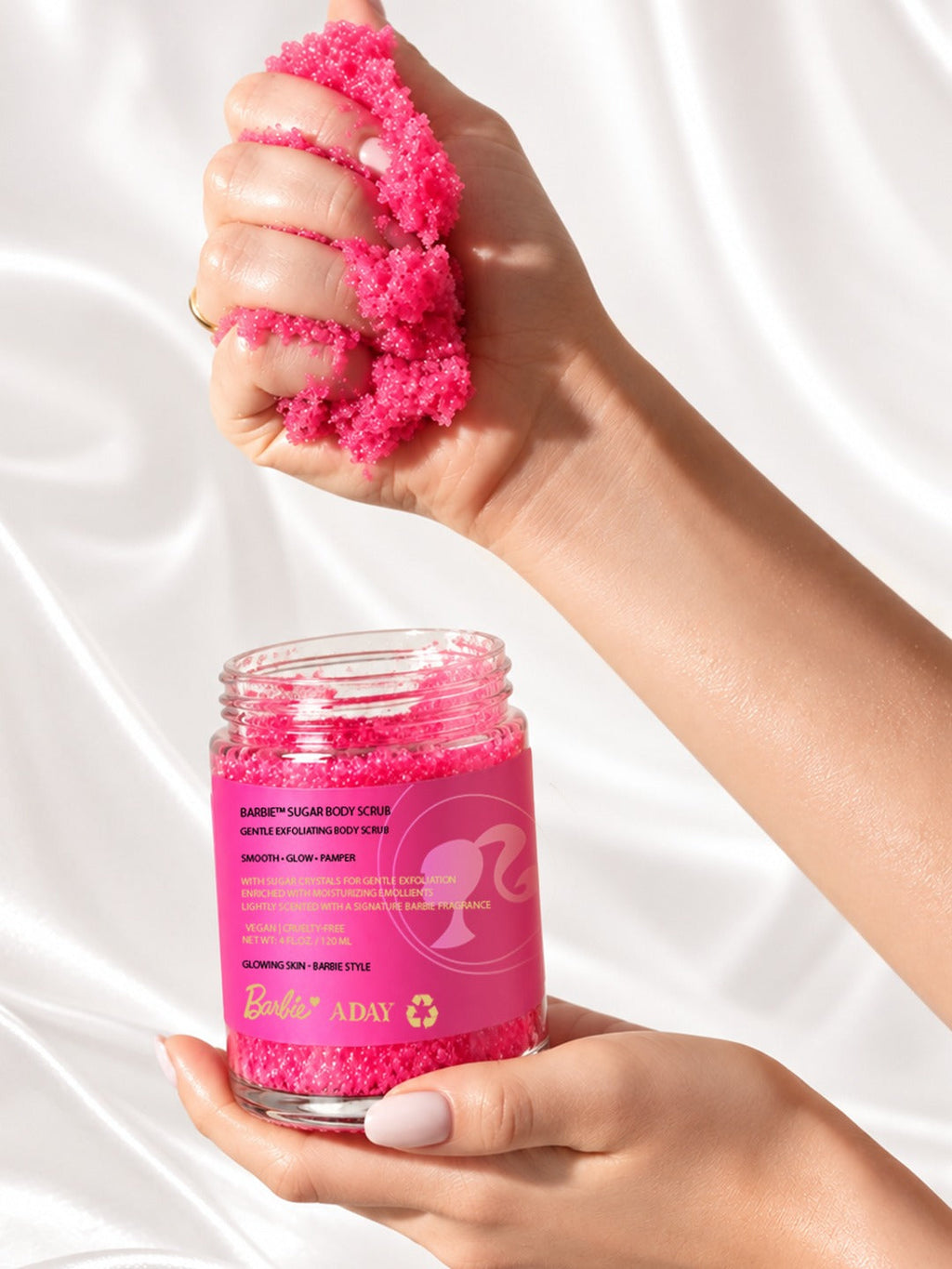 A hand lifting a scoop of ADAY x Barbie Sugar Body Scrub above the open jar
