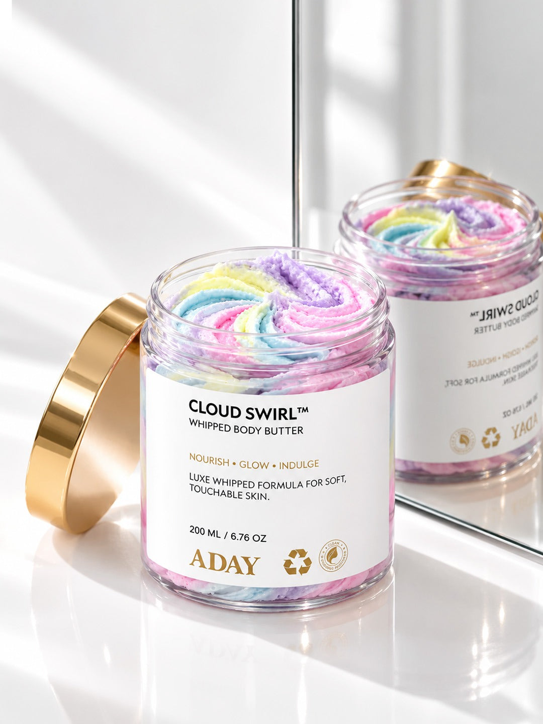 Open jar of ADAY Cloud Swirl Whipped Body Butter showing its pastel rainbow swirl, gold lid resting beside it
