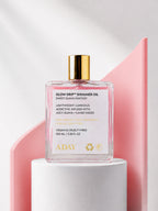 ADAY Glow Drip Shimmer Oil in Sweet Guava Fantasy, a 100 ml gold-capped glass bottle on a white pedestal