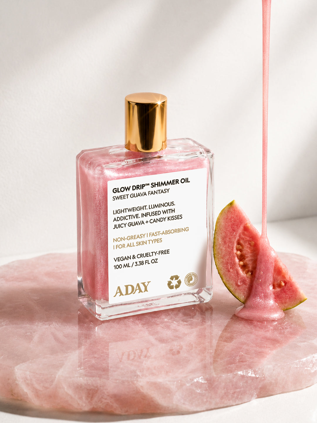 ADAY Glow Drip Shimmer Oil resting on blush silk beside a wedge of fresh guava
