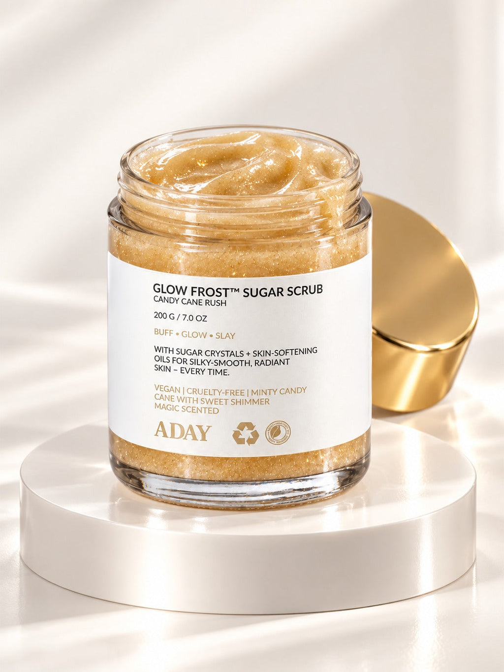 ADAY Glow Frost Sugar Scrub in Candy Cane Rush, a 200 g glass jar with a gold lid on a white pedestal