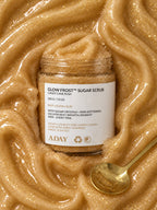 ADAY Glow Frost Sugar Scrub set in a swirl of golden scrub with a gold spoon
