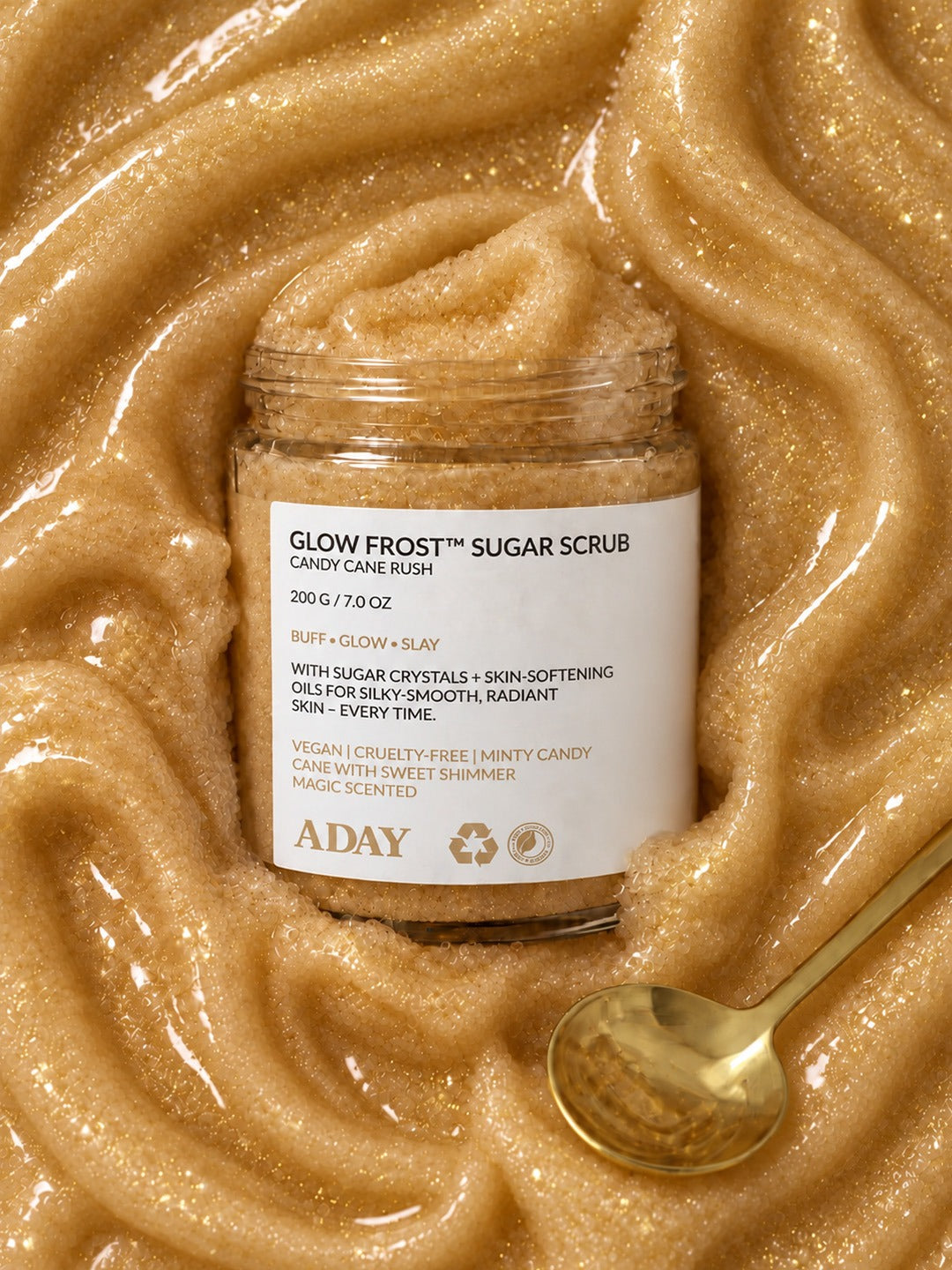 ADAY Glow Frost Sugar Scrub set in a swirl of golden scrub with a gold spoon