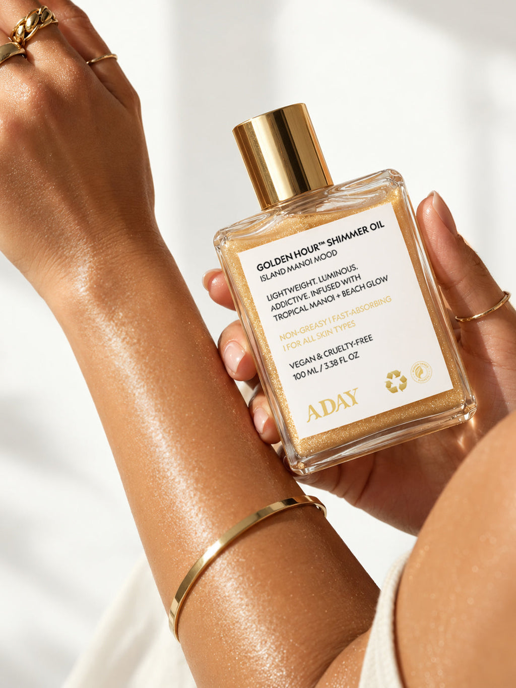 A hand wearing gold jewellery holding the ADAY Golden Hour Shimmer Oil