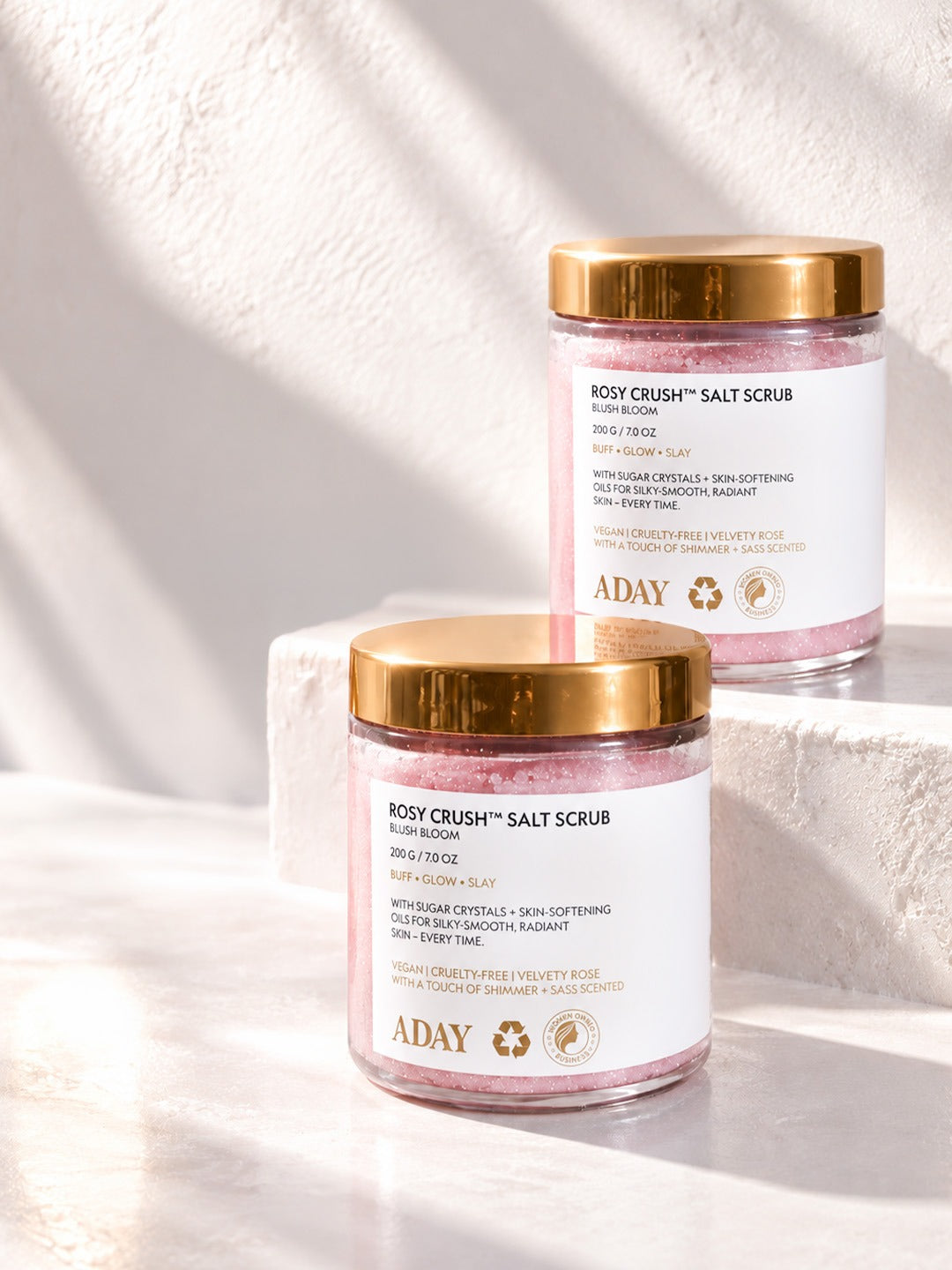 Two jars of ADAY Rosy Crush Salt Scrub on white plinths in sunlight