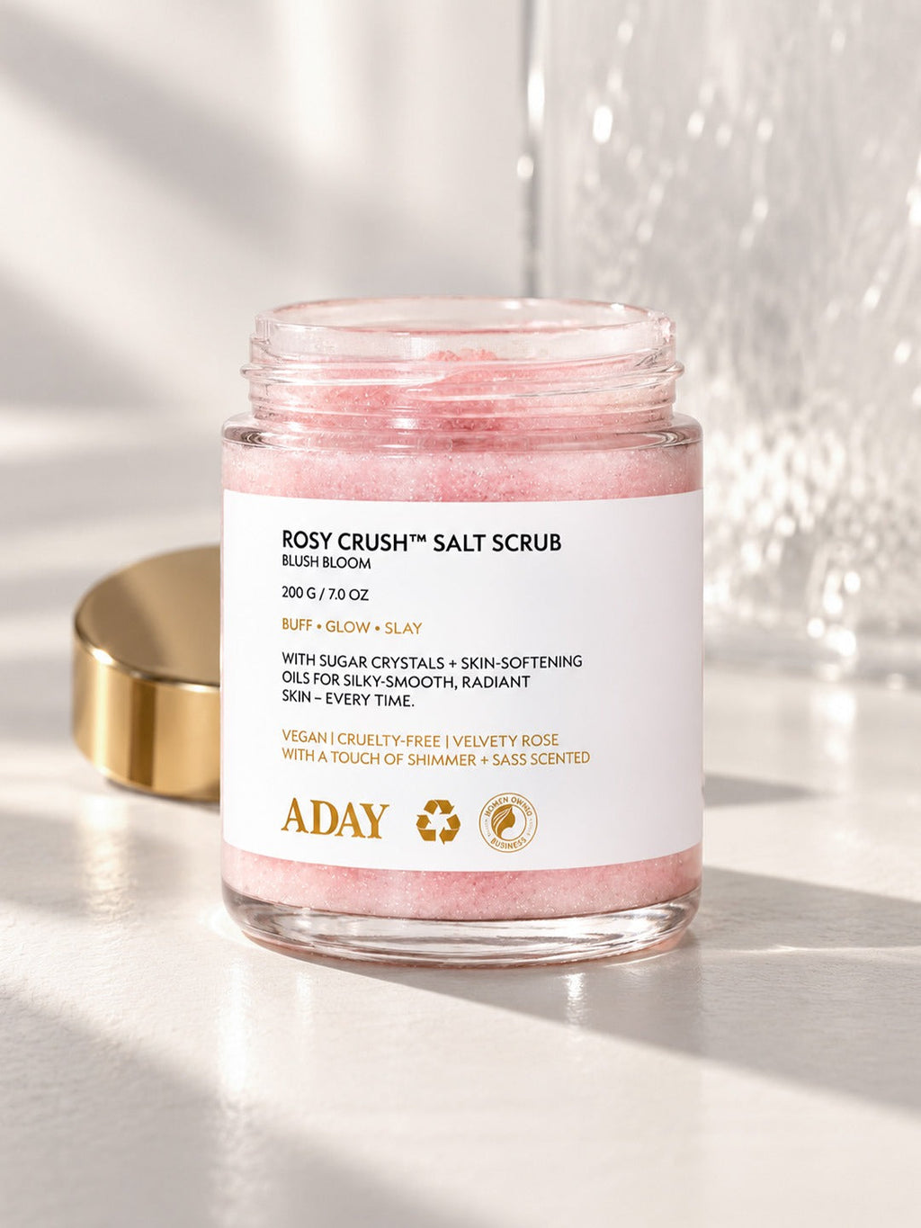 An open jar of ADAY Rosy Crush Salt Scrub in sunlight, gold lid set to one side