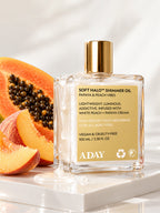 ADAY Soft Halo Shimmer Oil in Papaya & Peach Vibes, a 100 ml gold-capped glass bottle on a white plinth with papaya and peach slices