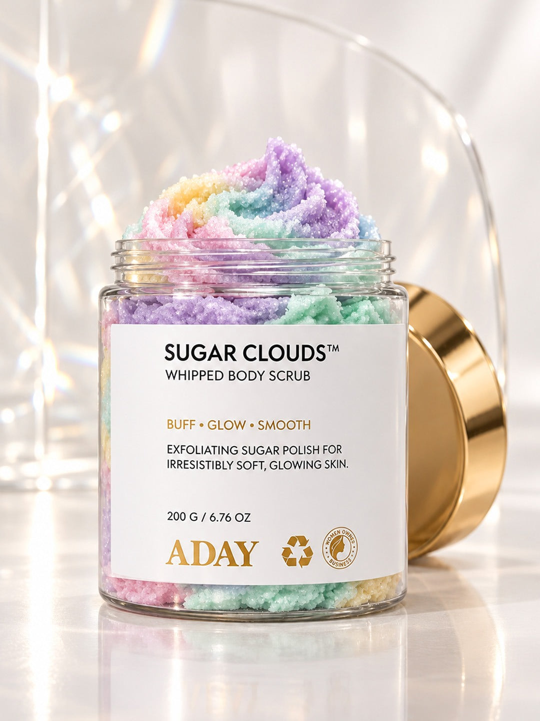 Open jar of ADAY Sugar Clouds Whipped Body Scrub with its pastel rainbow swirl peak, gold lid leaning against it